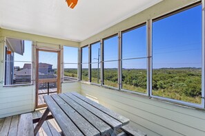 A covered patio with a wooden bench, large windows offering a view of greenery, and a door with a mesh screen.