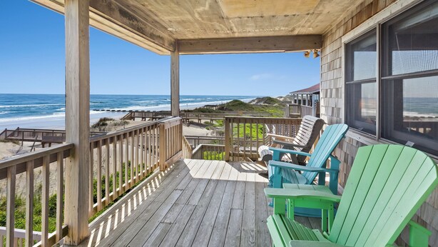 A wooden deck with a view of the ocean, featuring a railing and several Adirondack chairs.
