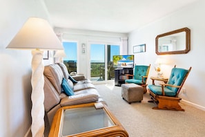 A living room with a sofa, armchair, and ottoman, a television, and a balcony with a view.