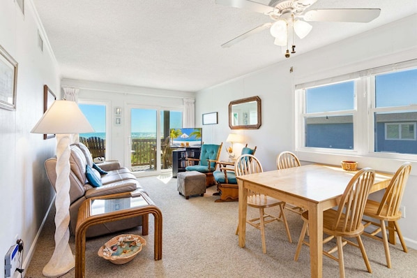 A spacious living room with a large window, a ceiling fan, and a dining area with wooden furniture.