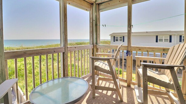Eine Holzterrasse mit Glastisch und Stühlen, mit Blick auf einen Strand und ein Gebäude.
