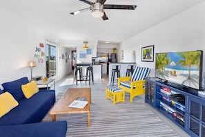 A modern living room with a sofa, coffee table, and a TV on a cabinet. There is a kitchen in the background with a dining area.