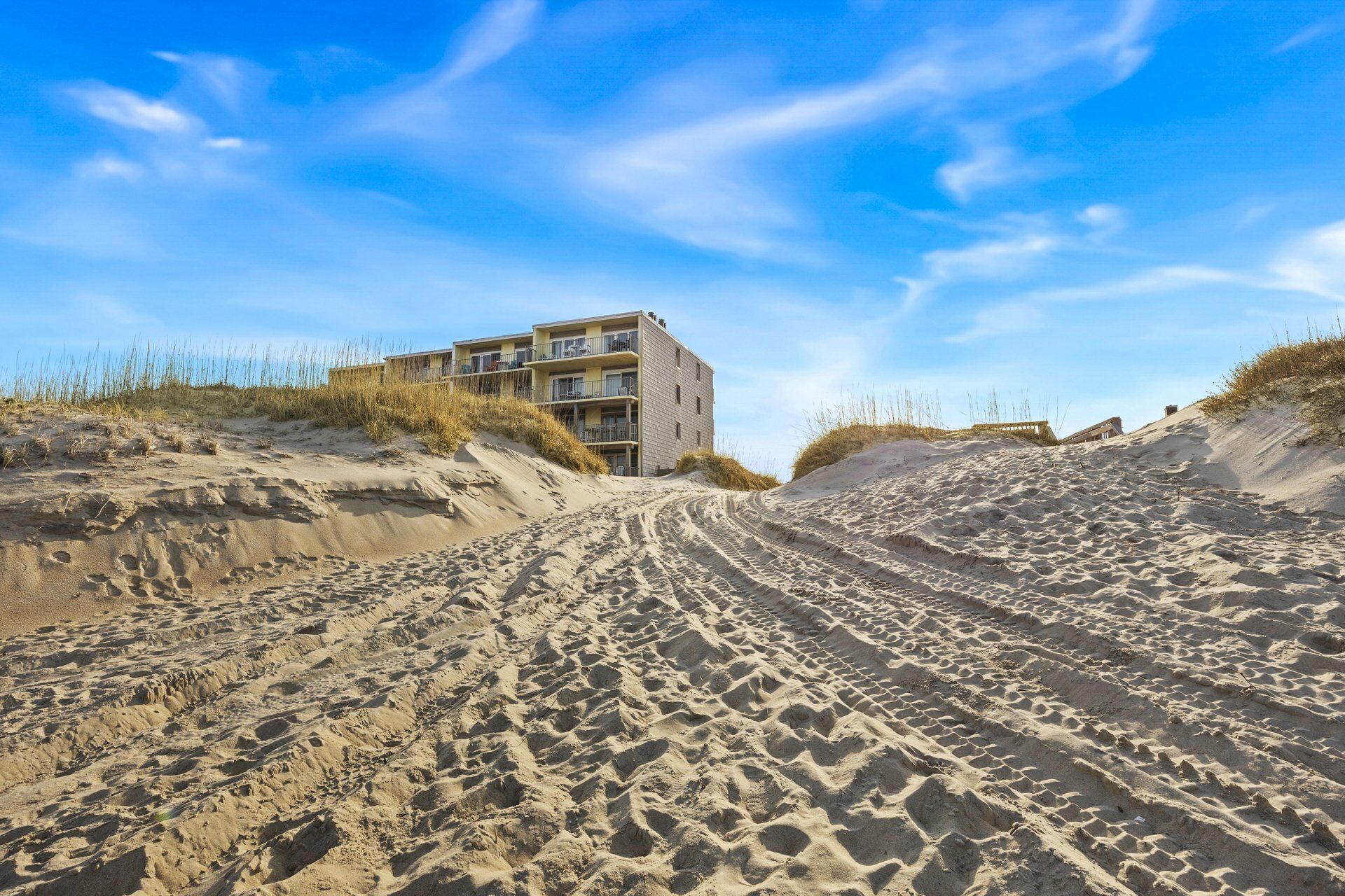 A beachfront building with multiple balconies, situated on sandy dunes with sparse vegetation.
