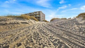 Un edificio frente a la playa con varios balcones, ubicado sobre dunas arenosas y vegetación escasa.