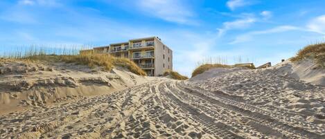 A beachfront building with multiple balconies, situated on sandy dunes with sparse vegetation.