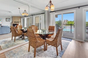 A dining area with wicker chairs and a glass table, a living room with a sofa and a fireplace, and a view of a pool through large sliding doors. - Oceanfront 2BR condo: pool, Jacuzzi & oceanfront (Nags Head)