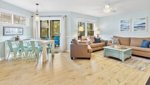 A spacious living room with a dining area, a ceiling fan, and large windows. - Cozy 2BR Retreat | Pool, Tennis & Quick Beach Access (Kill Devil Hills)