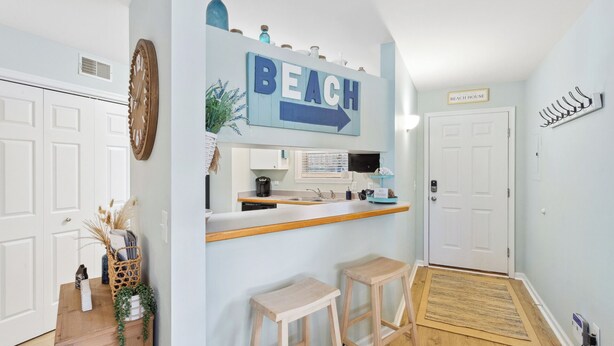 A kitchen with a 'BEACH' sign, wooden stools, and a small bar area. - Cozy 2BR Retreat | Pool, Tennis & Quick Beach Access (Kill Devil Hills)