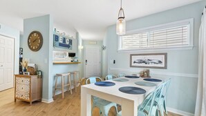A dining area with a white table set for six, light blue chairs, and a large window with blinds. - Cozy 2BR Retreat | Pool, Tennis & Quick Beach Access (Kill Devil Hills)