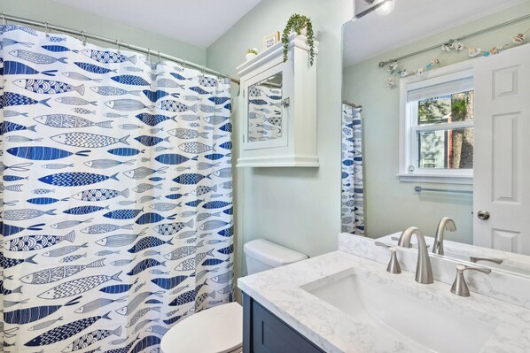 A bathroom with a shower curtain featuring a blue and white fish pattern, a white toilet, a marble sink with three faucets, and a mirror above the sink. - Cozy 2BR Retreat | Pool, Tennis & Quick Beach Access (Kill Devil Hills)