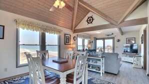 A spacious living area with a wooden ceiling, a dining table with chairs, a sofa, and a TV. Large windows offer a view of the beach. - Luxury Oceanfront Penthouse w/ Stunning Views (Kill Devil Hills)