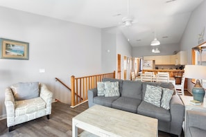 A living room with a grey sofa, patterned armchair, and a wooden coffee table. A staircase leads to a kitchen area with a dining table and chairs.