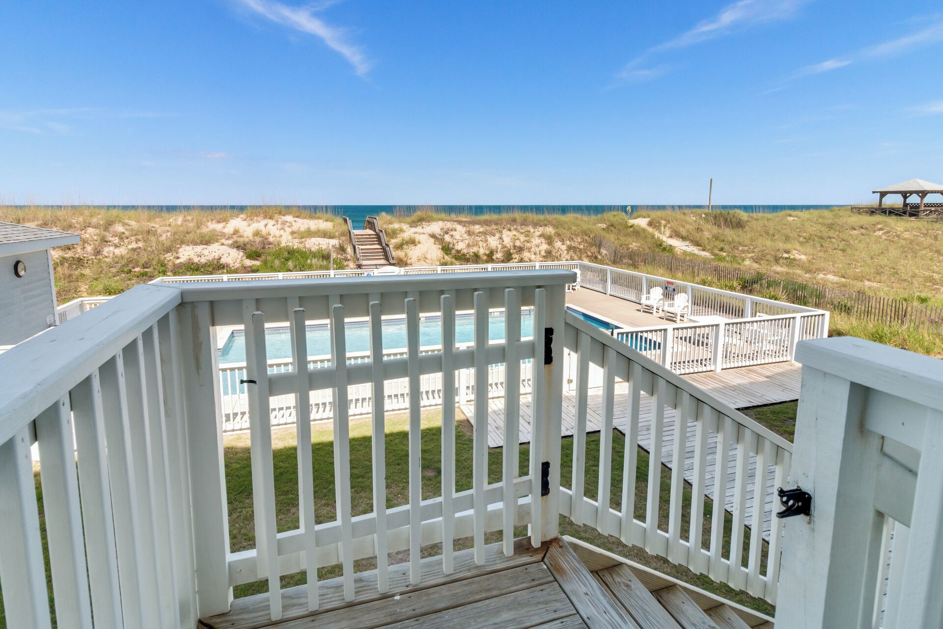 A wooden deck with a pool and outdoor seating, overlooking a beach and dunes.