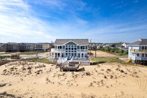 A beachfront property with a large house featuring multiple balconies, a sandy beach in the foreground, and other residential buildings in the background. - Fragile Magic: Oceanfront w/ Hot Tub & Private Walkway (Kitty Hawk)