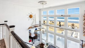 Una habitación amplia con techo alto, grandes ventanales con vistas a la playa, un comedor con mesa y sillas de madera, y una escalera con barandilla oscura.
