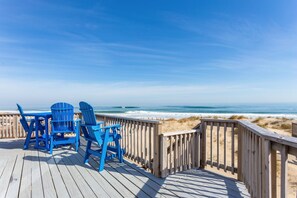 Terraza de madera con sillones Adirondack azules que dan a una playa.