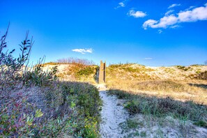 A pathway leading to a structure in a sandy, vegetated area with a clear sky.