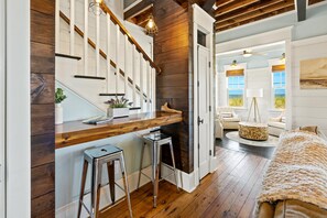 A room with a wooden staircase, a kitchen area with stools, a dining area with a table and chairs, and a bed with a textured blanket.