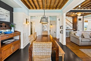 A spacious dining area with a wooden table and wicker chairs, a kitchen with white cabinetry, and a living area with a sofa and a view of the outdoors.