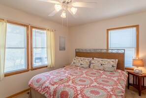 Une chambre à coucher avec un lit, un ventilateur de plafond et deux fenêtres équipées de stores.