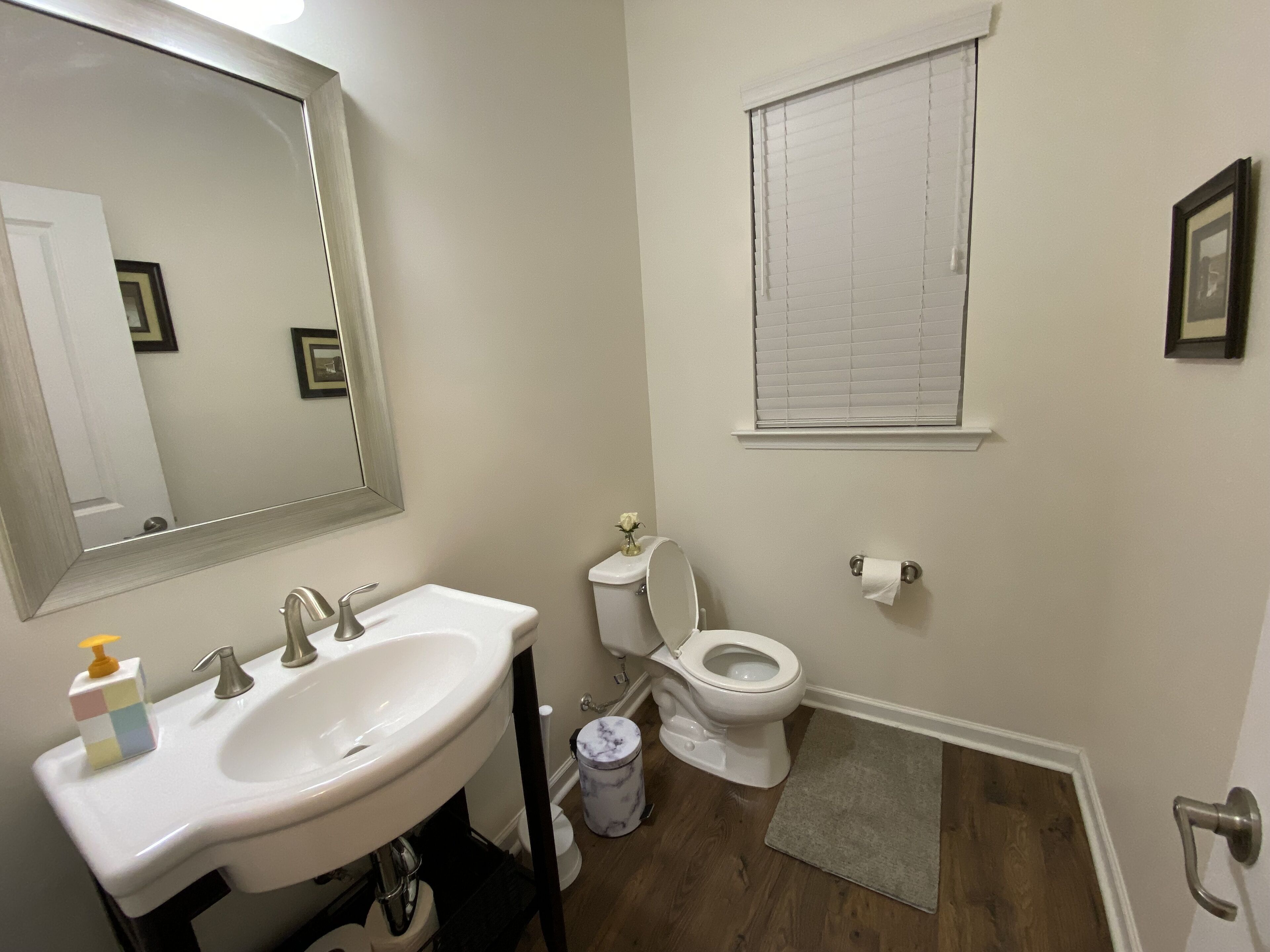 Un baño con lavamanos blanco, espejo, inodoro y ventana con persianas.