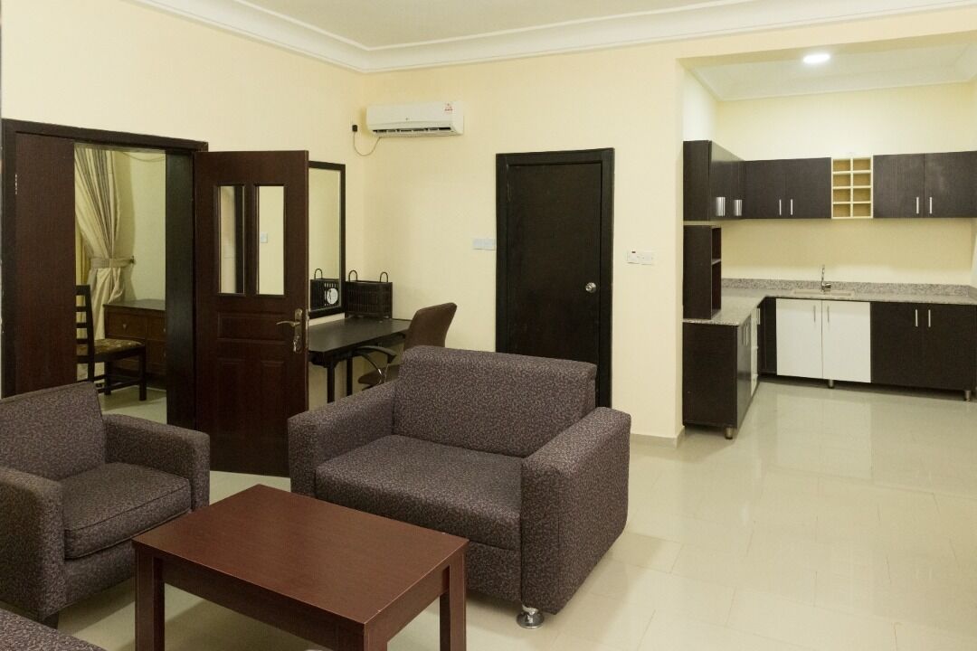 Un espace de vie compact comprenant un coin cuisine, deux fauteuils, une petite table et une porte donnant sur une autre pièce.