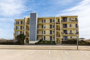A multi-story residential building with balconies, a central stairwell, and a parking lot in front. - Beachfront OBX condo: family fun & ocean views (Nags Head)