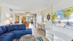 A living room with a blue sofa, a dining area, a kitchen, and a large TV displaying a beach scene. - Beachfront OBX condo: family fun & ocean views (Nags Head)