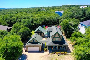 Une grande maison à plusieurs étages, dotée d’un garage, entourée d’une végétation luxuriante et bordée d’autres maisons au loin.