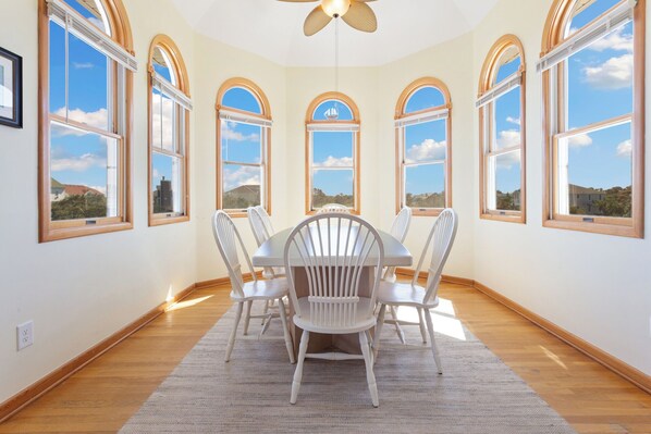 Enjoy meals in this spacious dining area with a round table and six chairs, surrounded by large windows offering a view of the sky and distant landscape.