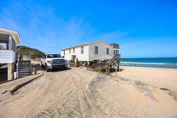 Una casa de playa con un vehículo estacionado adelante, escaleras de madera que llevan a la entrada y una vista despejada del océano.