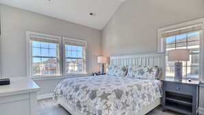 Une chambre avec un grand lit, deux fenêtres équipées de stores, une table de chevet avec une lampe et une commode avec une lampe.
