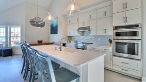 Une cuisine moderne avec des meubles blancs, un grand îlot central doté de tabourets, des appareils en acier inoxydable et un éclairage suspendu.