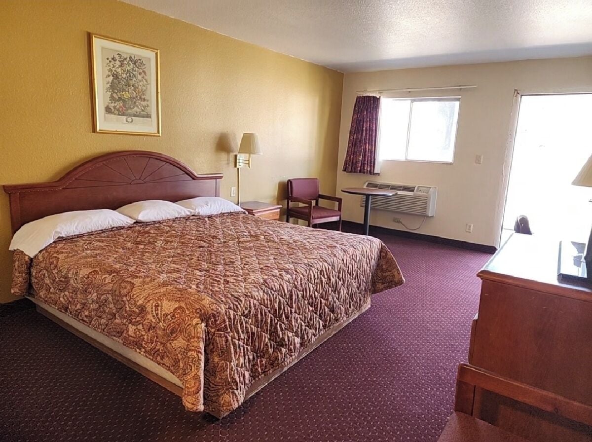 Un cuarto de hotel con una cama grande, un escritorio, una silla, un ventanal con cortinas, un equipo de aire acondicionado y un cuadro en la pared.