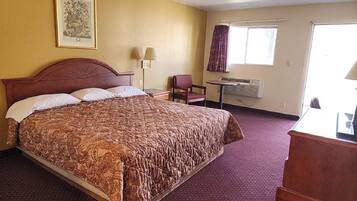Un cuarto de hotel con una cama grande, un escritorio, una silla, un ventanal con cortinas, un equipo de aire acondicionado y un cuadro en la pared.