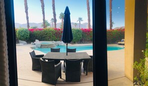 A pool area with a hot tub, surrounded by palm trees and red flowers. There are wicker chairs and a blue umbrella.