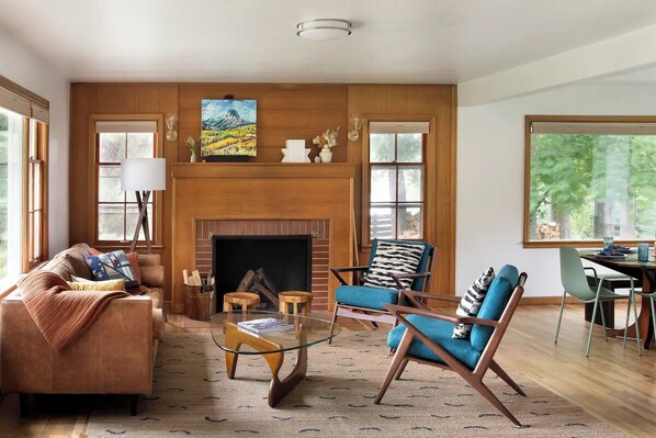 A spacious living room with a fireplace, wooden paneling, a sofa, armchairs, and a dining area with a table and chairs.