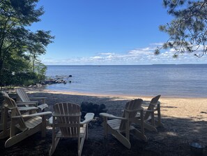 A beach with wooden chairs and a fire pit, overlooking a calm sea and distant shoreline.