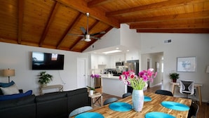 Ein modernes Wohnzimmer mit einem Holzesstisch, blauen Tischsets, einem schwarzen Sofa und einem an der Wand befestigten Fernseher.