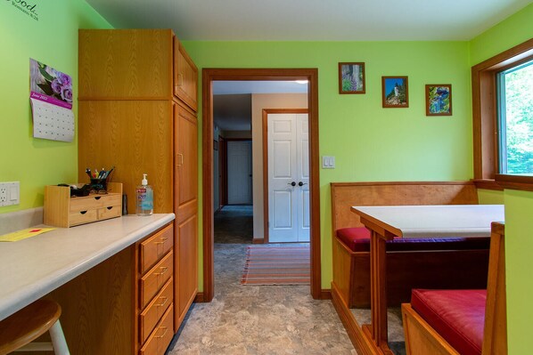 Una habitación pequeña y luminosa con un escritorio, un banco y una puerta que da a otra habitación.