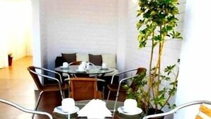 A round table with four chairs, a sofa, and a potted plant in a room with a wooden ceiling and white walls.
