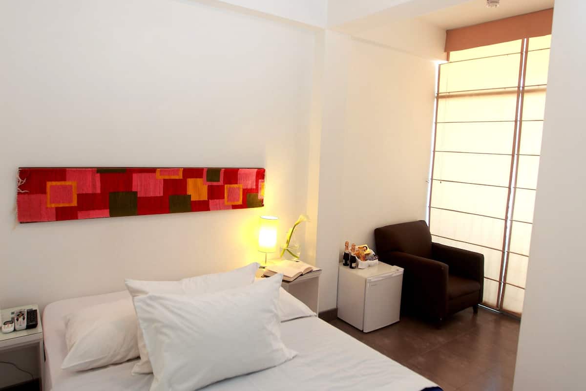 Ein Hotelzimmer mit Bett, Nachttisch, Lampe, Stuhl und einem Fenster mit Jalousien.