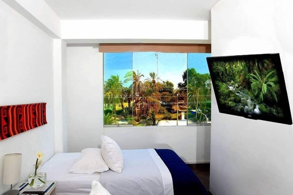 Ein modernes Hotelzimmer mit zwei Betten, einem großen Fenster mit Blick auf Palmen und einen Strand, sowie einem an der Wand befestigten Fernseher.