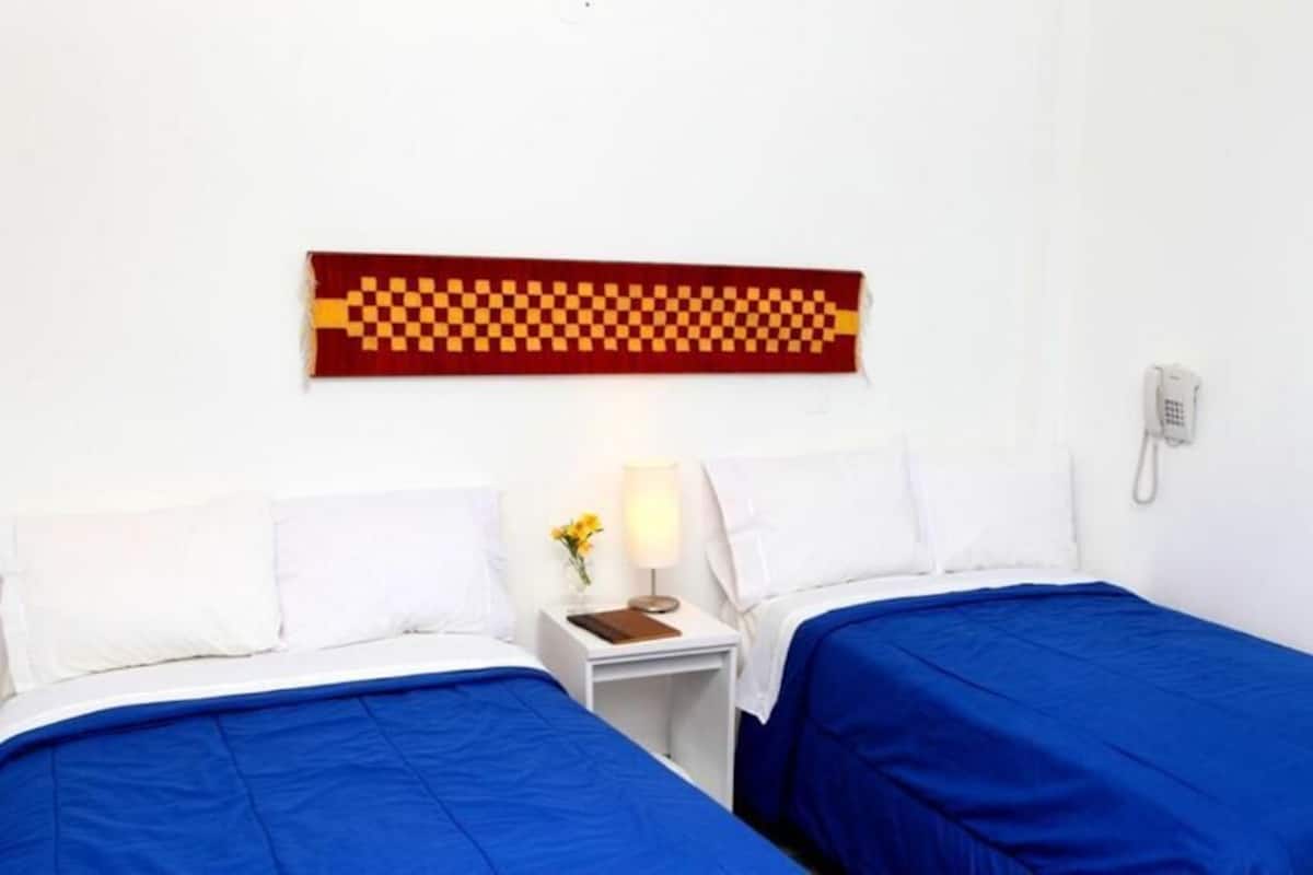 Ein Hotelzimmer mit zwei Betten, einem Nachttisch mit Lampe, einem Telefon und einem an der Wand befestigten Kunstwerk.