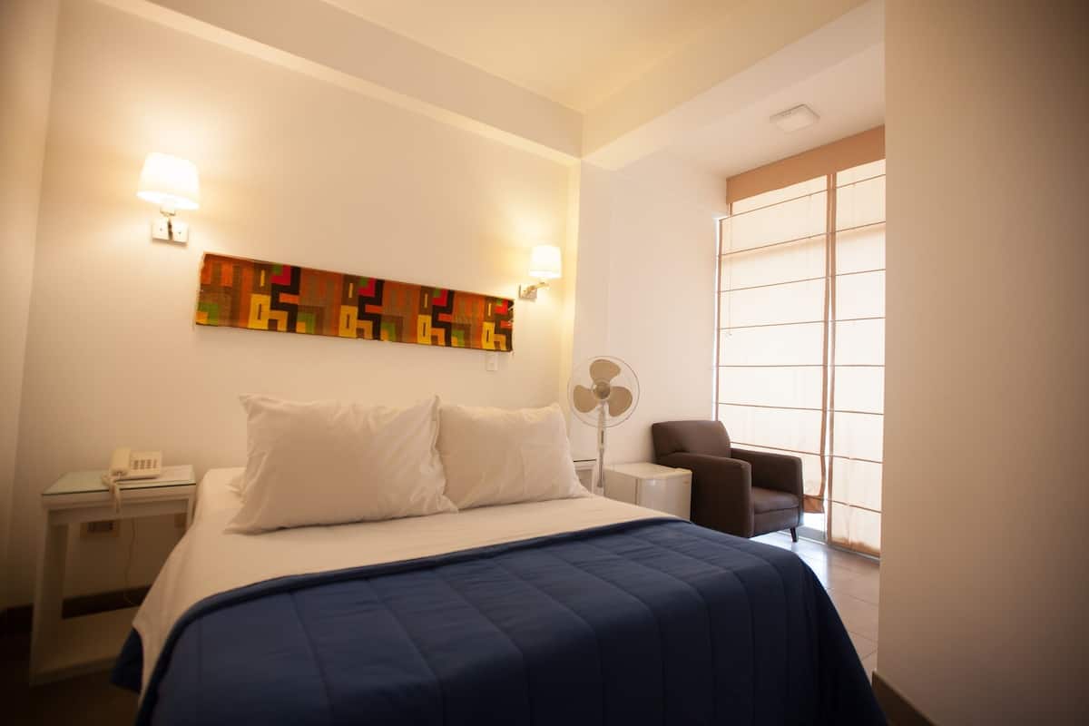 Ein Hotelzimmer mit einem Bett, einem Sessel, einem Schreibtisch mit Telefon, einem Ventilator und einem Fenster mit Jalousien.