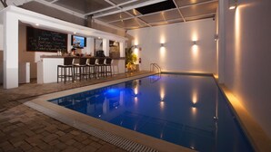 An indoor swimming pool with a bar area, seating, and a television screen.