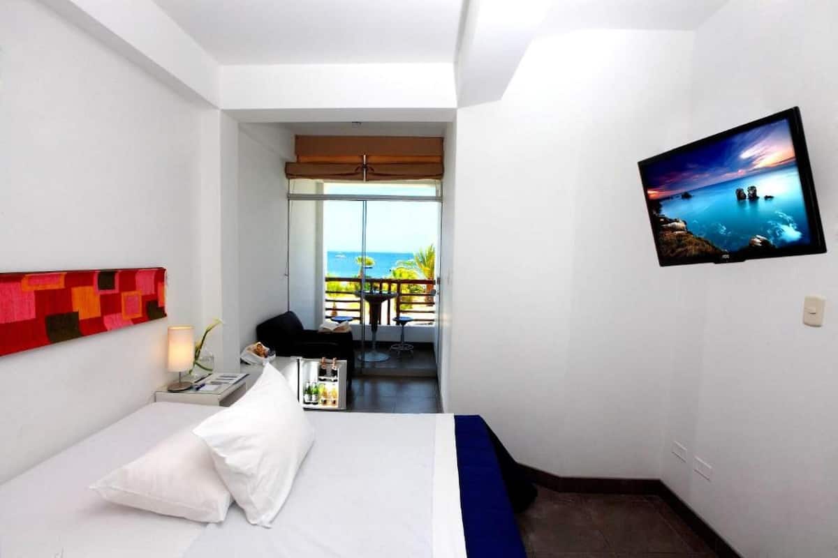 Ein Hotelzimmer mit Bett, Fernseher an der Wand, einem Balkon mit Meerblick und einem kleinen Tisch mit Lampe und Blumenvase.