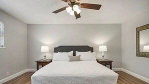 Une chambre à coucher avec un grand lit, deux tables de chevet avec des lampes, un ventilateur de plafond et un miroir au mur.