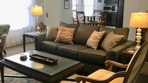 Un salon avec un canapé, une table basse et des fauteuils. On y trouve un ventilateur de plafond, un coin repas à l’arrière-plan et une cuisine visible à travers une porte ouverte.