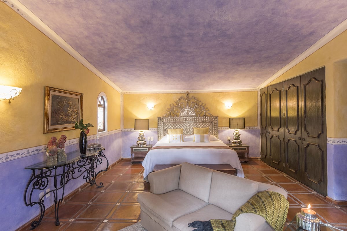 A bedroom with a bed, a sofa, a nightstand, a table with decorative items, and a wall-mounted lamp.